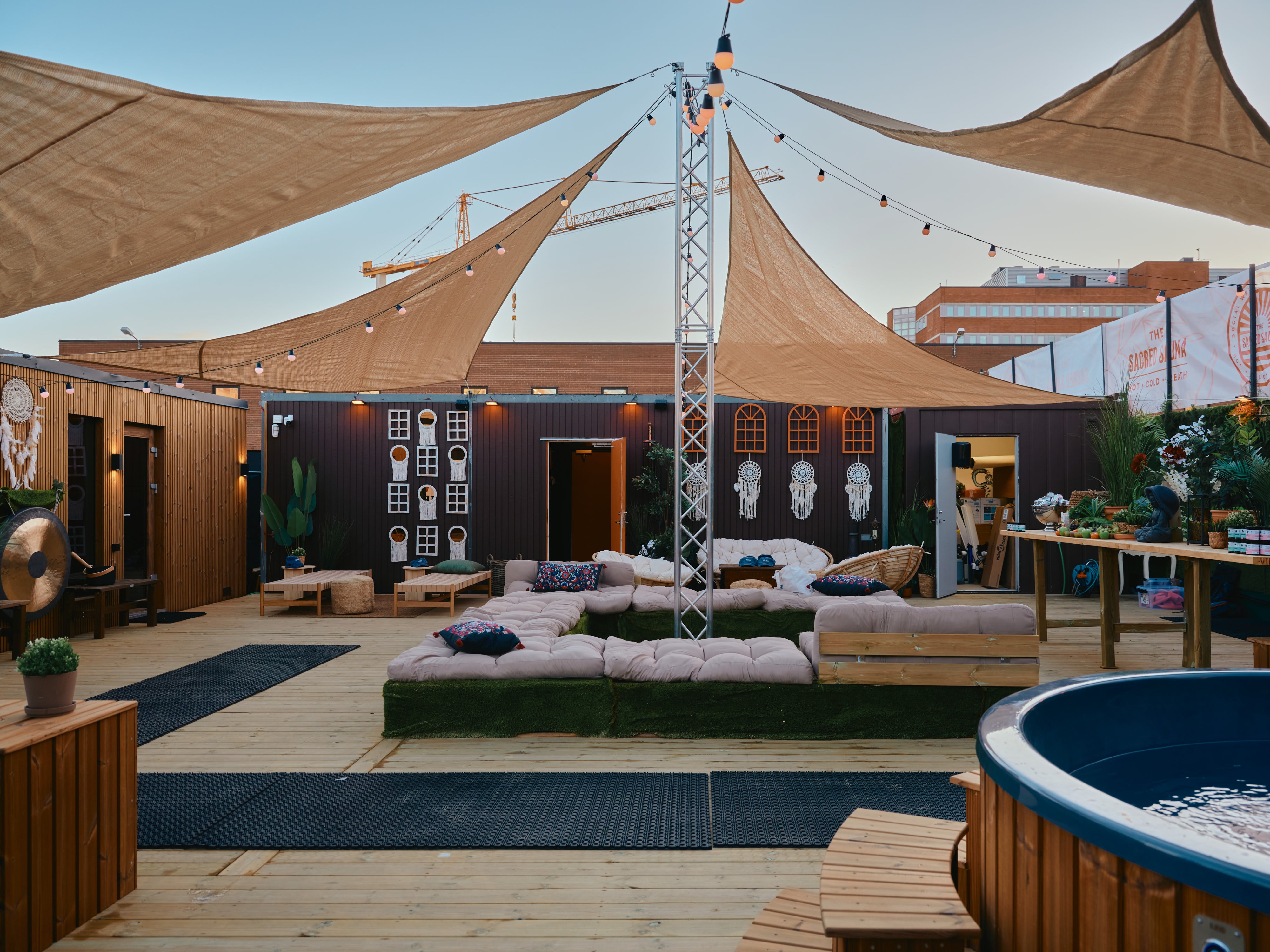 Upplyst utomhusområde med komfortabla sittplatser, träpaneler och dekorativa detaljer som drömfångare och en gonggong. Tältliknande strukturer och ljusslingor skapar en avslappnad atmosfär för sociala sammankomster.