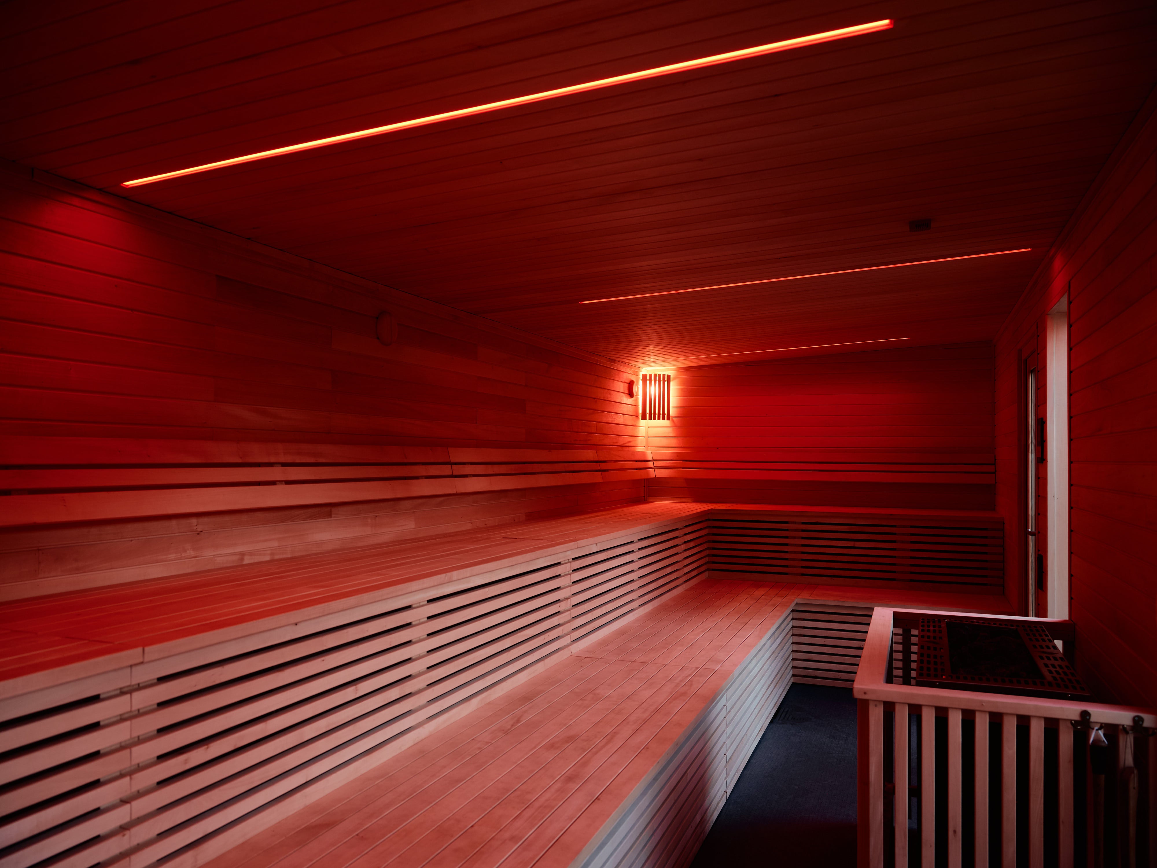 Inre vy av en modern bastu med röd belysning. Bastun har träpaneler, bänkar och en ventilationsenhet. Den röda ljussättningen skapar en avkopplande och atmosfärisk miljö.