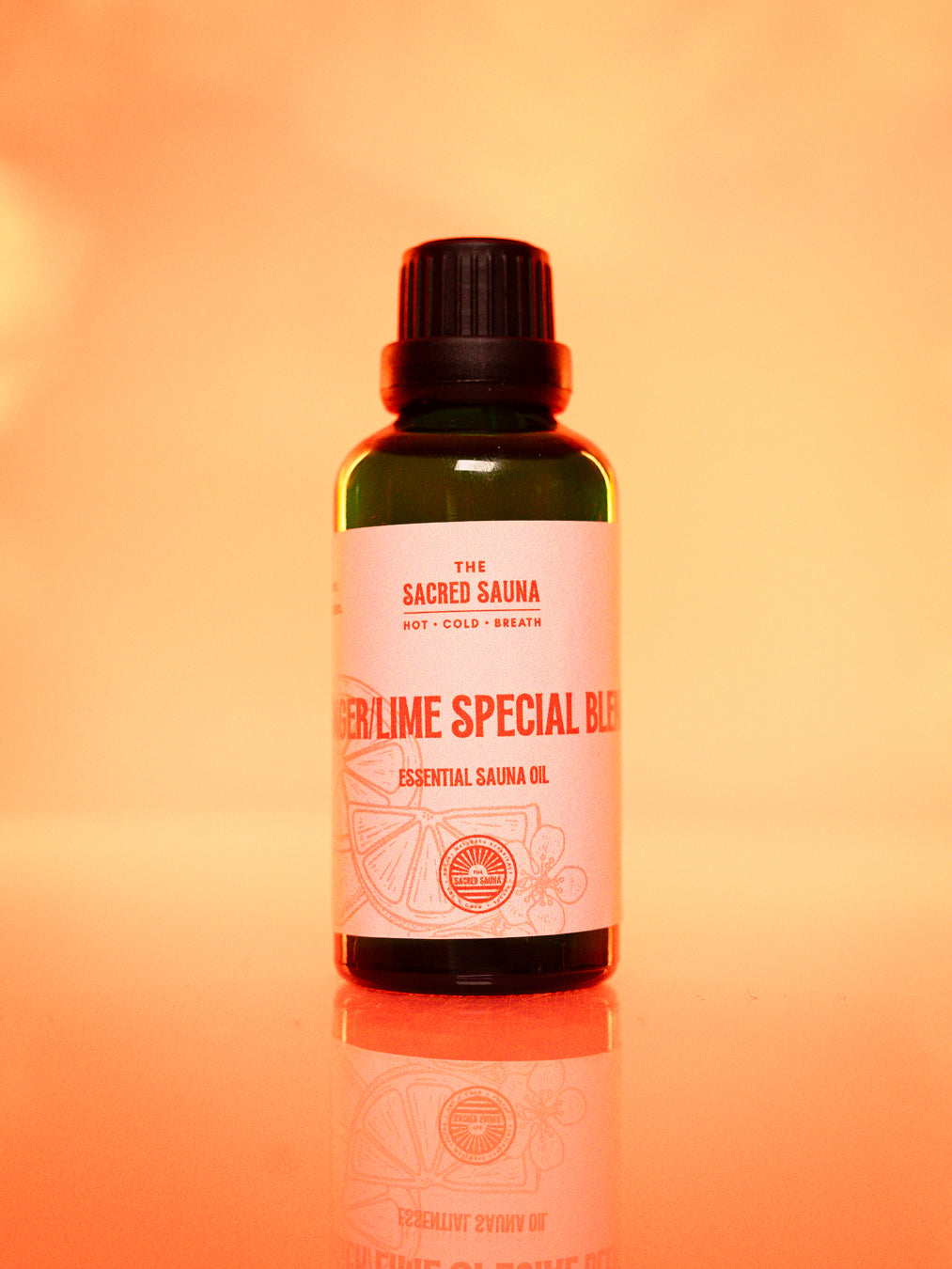 Flaska med 'Ginger/Lime Special Blend' eterisk olja från The Sacred Sauna, stående på en reflekterande yta med mjuk orange bakgrund. Flaskan är designad för användning i bastu och ger en uppfriskande doft av ingefära och lime.
