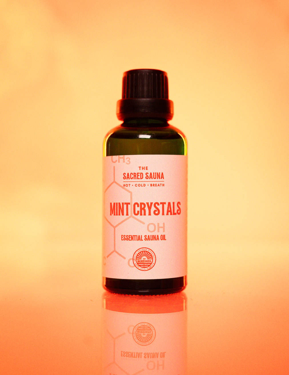 Flaska med 'Mint Crystals' eterisk olja från The Sacred Sauna, stående på en reflekterande yta med mjuk orange bakgrund. Flaskan är designad för användning i bastu och ger en uppfriskande mintdoft.