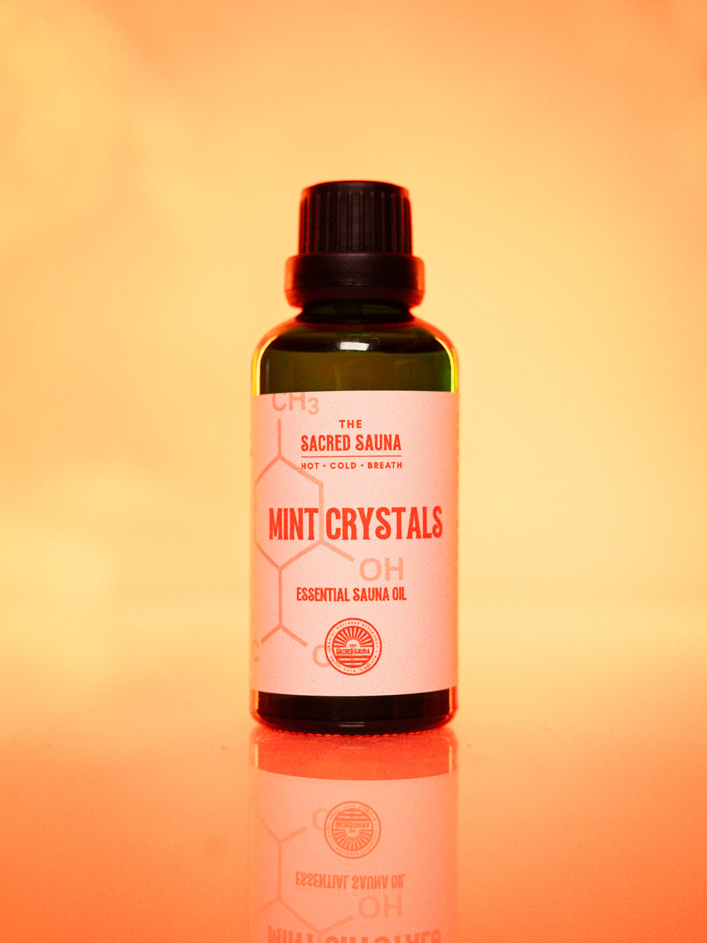 Flaska med 'Mint Crystals' eterisk olja från The Sacred Sauna, stående på en reflekterande yta med mjuk orange bakgrund. Flaskan är designad för användning i bastu och ger en uppfriskande mintdoft.