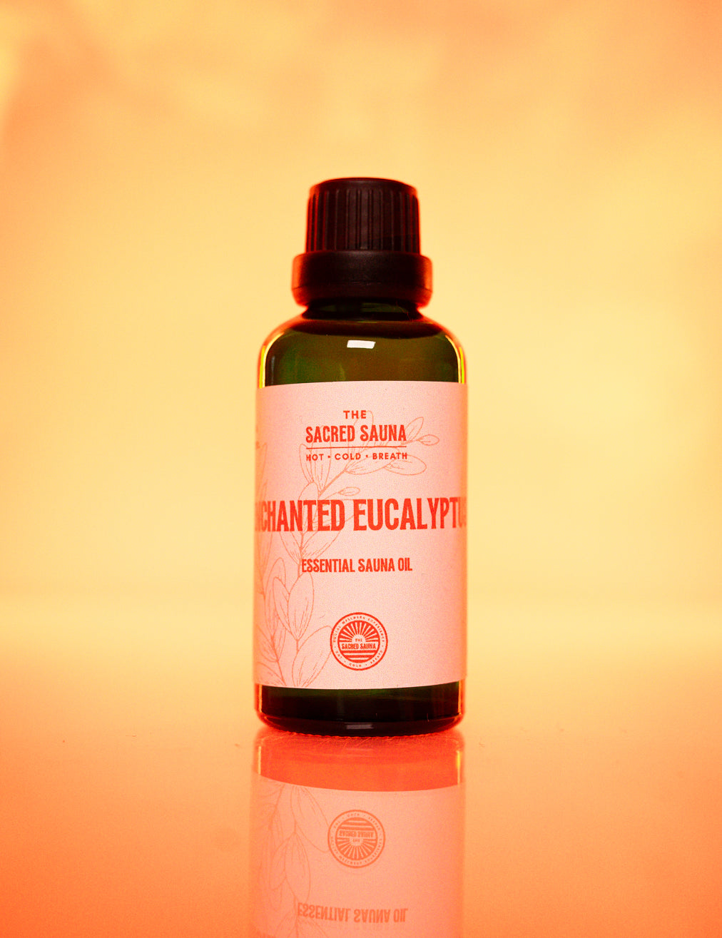 Flaska med 'Enchanted Eucalyptus' eterisk olja från The Sacred Sauna, stående på en reflekterande yta med mjuk orange bakgrund. Flaskan är designad för användning i bastu och ger en lugnande eukalyptusdoft.