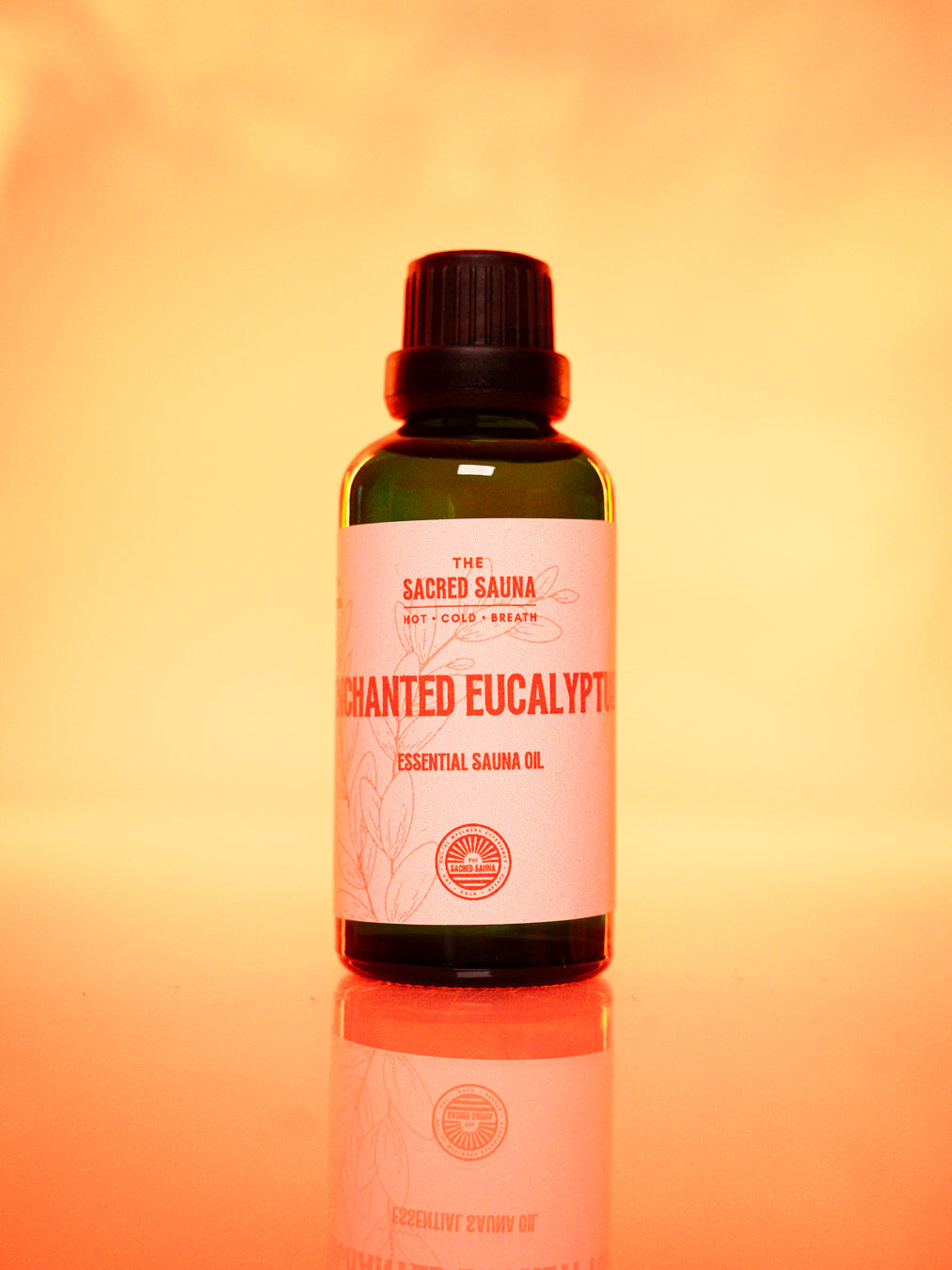 Flaska med 'Enchanted Eucalyptus' eterisk olja från The Sacred Sauna, stående på en reflekterande yta med mjuk orange bakgrund. Flaskan är designad för användning i bastu och ger en lugnande eukalyptusdoft.