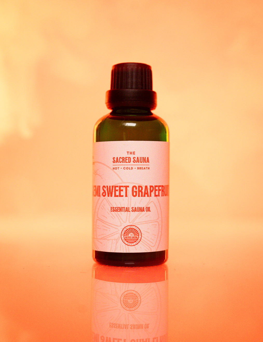 Flaska med 'Semi Sweet Grapefruit' eterisk olja från The Sacred Sauna, stående på en reflekterande yta med mjuk orange bakgrund. Flaskan är designad för användning i bastu och ger en uppfriskande citrusdoft.
