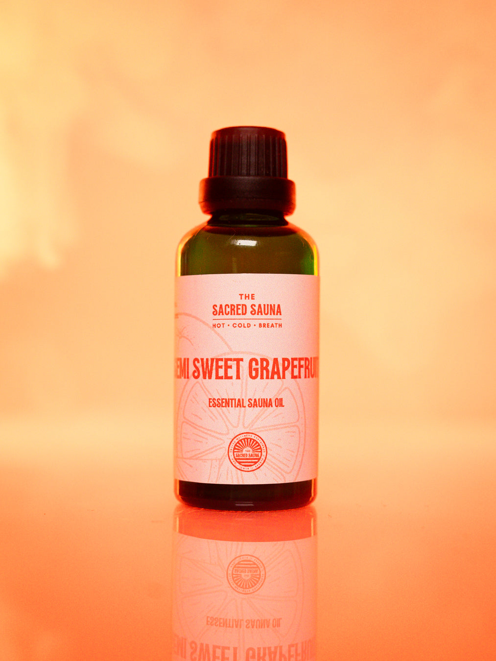 Flaska med 'Semi Sweet Grapefruit' eterisk olja från The Sacred Sauna, stående på en reflekterande yta med mjuk orange bakgrund. Flaskan är designad för användning i bastu och ger en uppfriskande citrusdoft.