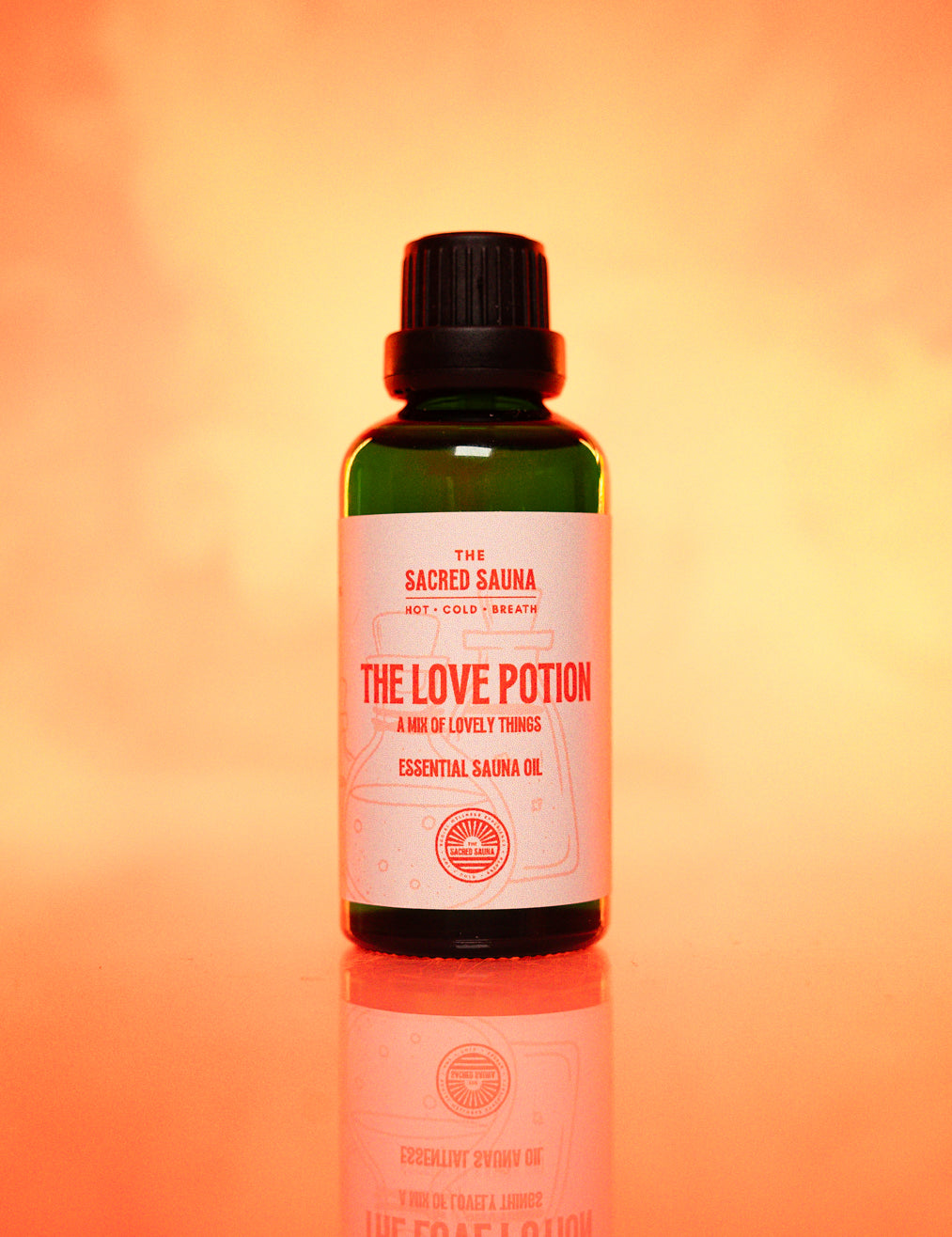 Flaska med 'The Love Potion' eterisk olja från The Sacred Sauna, stående på en reflekterande yta med mjuk orange bakgrund. Flaskan är designad för användning i bastu och erbjuder en blandning av härliga dofter för att skapa en romantisk atmosfär.
