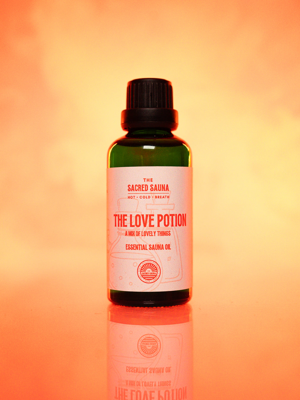 Flaska med 'The Love Potion' eterisk olja från The Sacred Sauna, stående på en reflekterande yta med mjuk orange bakgrund. Flaskan är designad för användning i bastu och erbjuder en blandning av härliga dofter för att skapa en romantisk atmosfär.