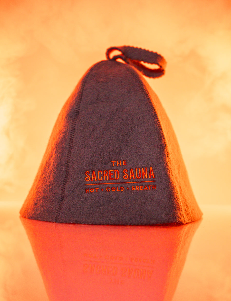 En grå bastumössa med logotypen 'The Sacred Sauna' i orange text. Mössan står på en reflekterande yta med en mjuk orange bakgrund, vilket skapar en varm och inbjudande atmosfär.