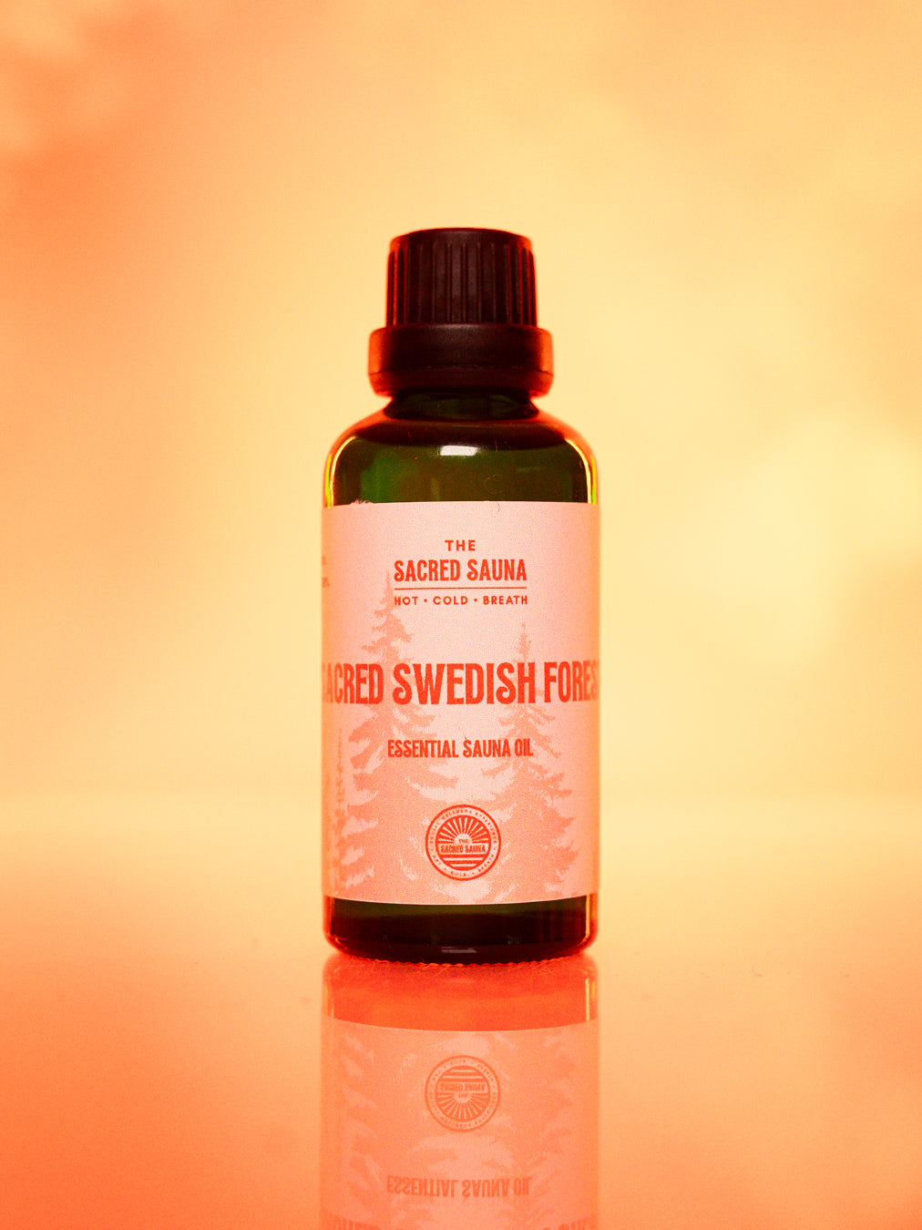 Flaska med 'Sacred Swedish Forest' eterisk olja från The Sacred Sauna, stående på en reflekterande yta med mjuk orange bakgrund. Flaskan är designad för användning i bastu och ger en frisk doft av svenska skogar.