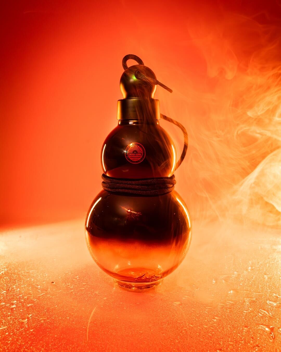 En vattenglasflaska formad som en traditionell termos, med The Sacred Sauna-logotyp på, omgiven av rök och orange belysning. Flaskan är bundet med ett rep och står på en fuktig yta, vilket skapar en dramatisk och mystisk atmosfär.
