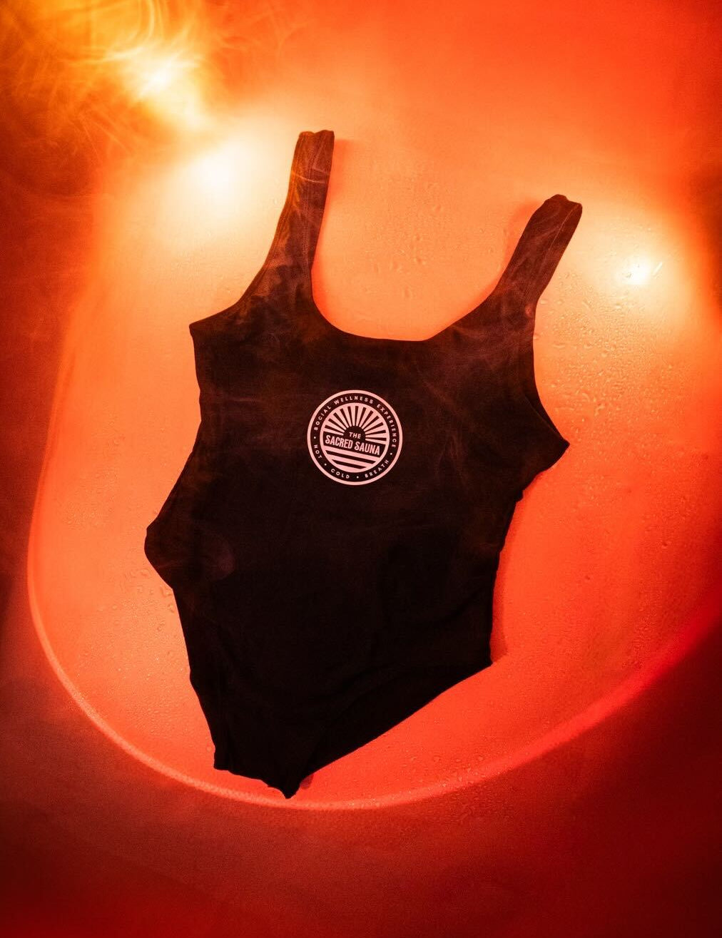 Svart baddräkt med The Sacred Sauna-logotyp på framsidan, hängande mot en fuktig yta med röd bakgrund och rök. Baddräkten är designad för bastubad och avkoppling.