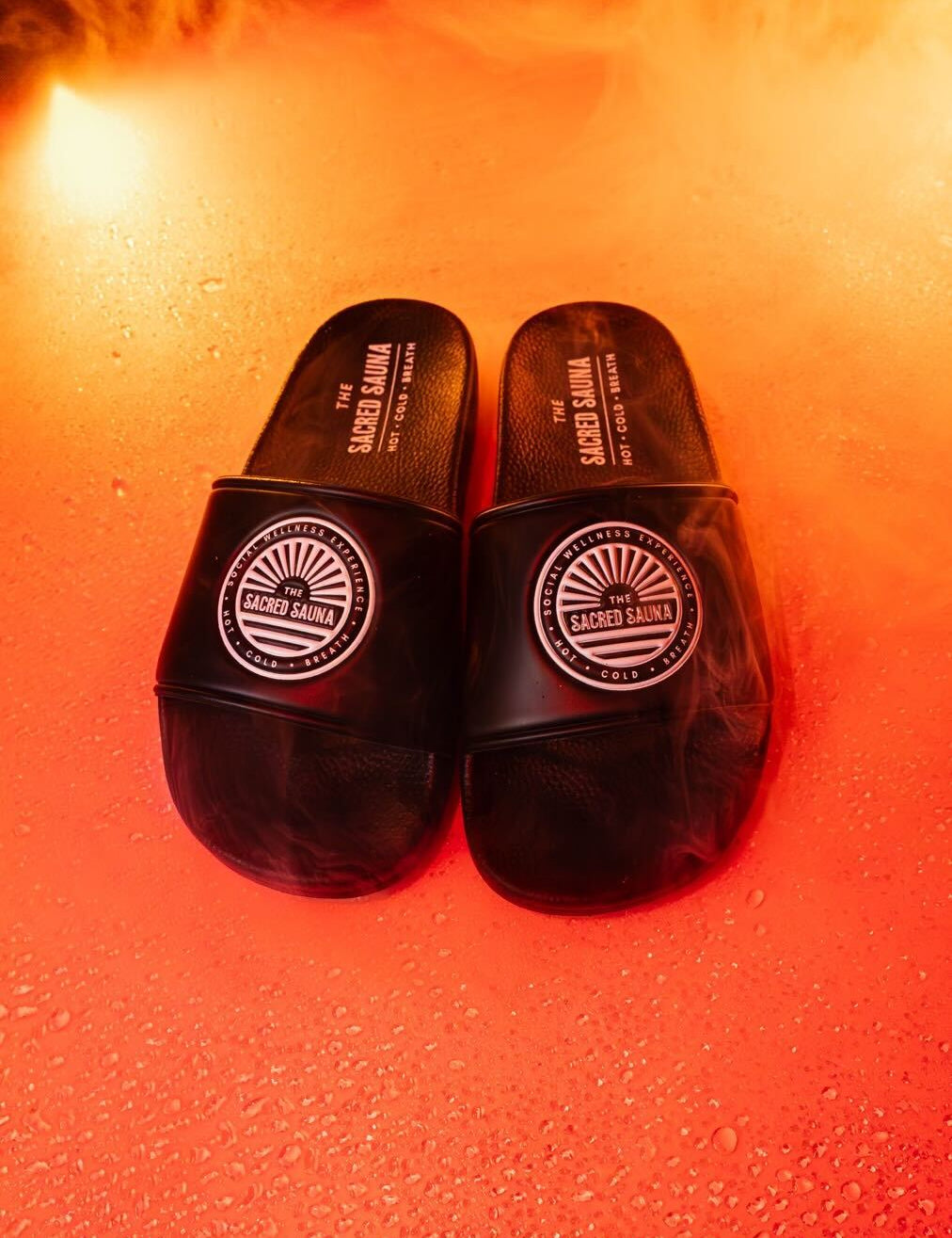 Svarta sandaler med The Sacred Sauna-logotyp på remmen, placerade på en fuktig yta med orange bakgrund och rök. Sandalerna är designade för användning vid bastu eller avkoppling och bär texten 'HOT · COLD'.