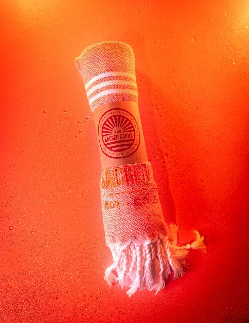 En handduk från The Sacred Sauna, rullad och med logotypen 'THE SACRED SAUNA' samt texten 'HOT · COLD' tryckt på den. Handduken ligger på en fuktig yta med orange bakgrund, vilket skapar en varm och inbjudande atmosfär.