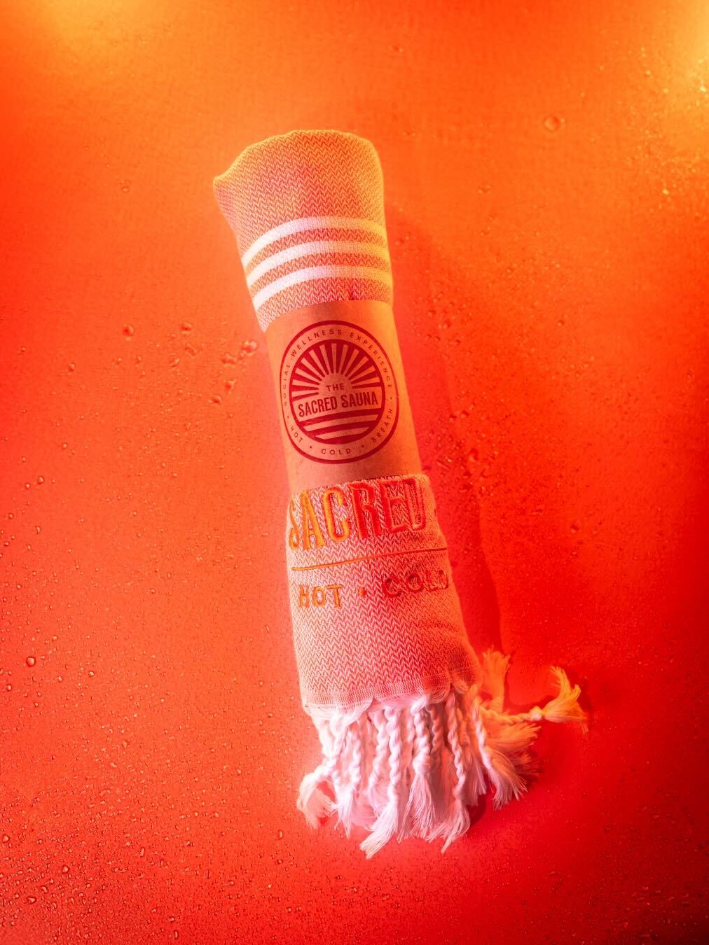 En handduk från The Sacred Sauna, rullad och med logotypen 'THE SACRED SAUNA' samt texten 'HOT · COLD' tryckt på den. Handduken ligger på en fuktig yta med orange bakgrund, vilket skapar en varm och inbjudande atmosfär.