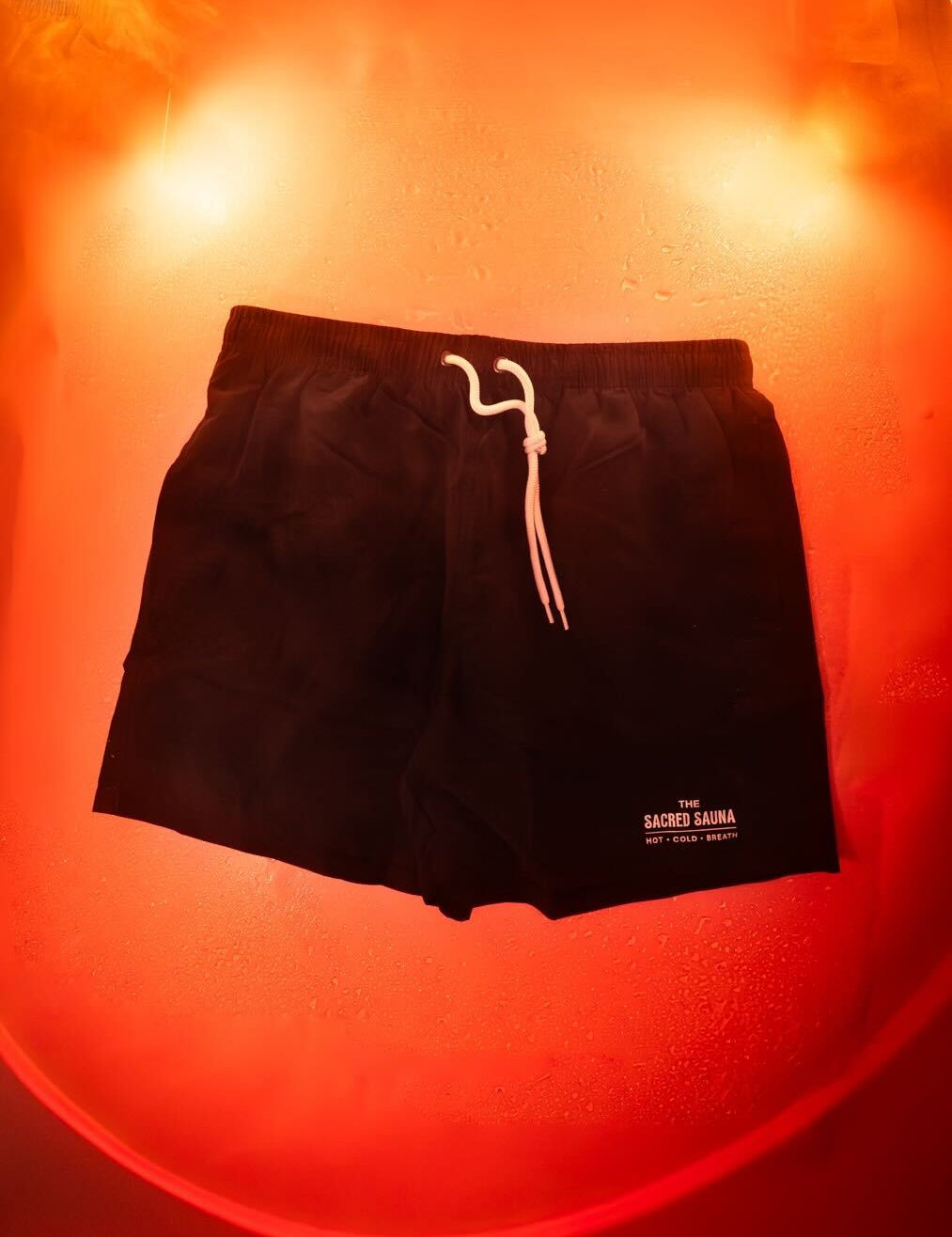 Svarta shorts med The Sacred Sauna-logotyp på fållen, hängande på en fuktig yta med röd bakgrund och rök. Shorts är designade för bastubad eller avkoppling.