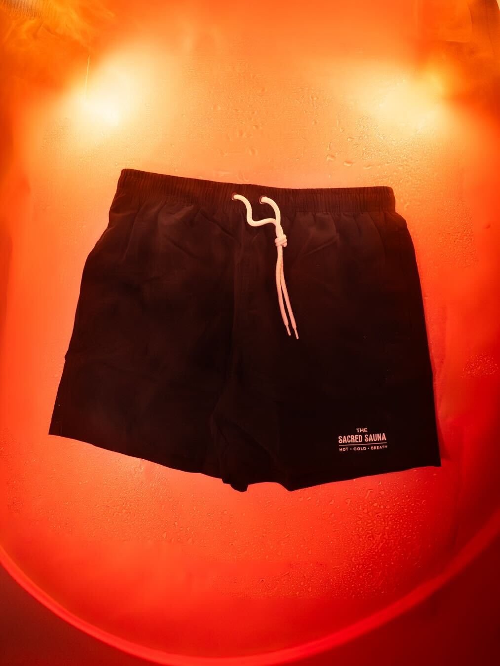 Svarta shorts med The Sacred Sauna-logotyp på fållen, hängande på en fuktig yta med röd bakgrund och rök. Shorts är designade för bastubad eller avkoppling.