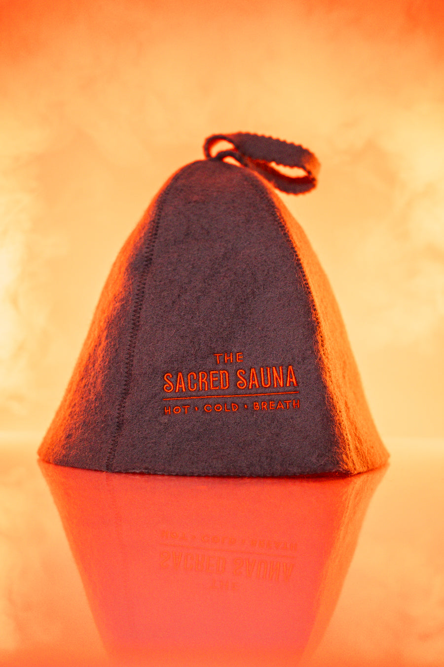 En grå bastumössa med logotypen &#39;The Sacred Sauna&#39; i orange text. Mössan står på en reflekterande yta med en mjuk orange bakgrund, vilket skapar en varm och inbjudande atmosfär.