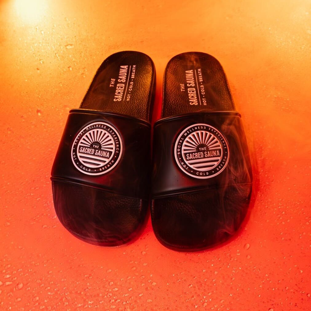 Svarta sandaler med The Sacred Sauna-logotyp på remmen, placerade på en fuktig yta med orange bakgrund och rök. Sandalerna är designade för användning vid bastu eller avkoppling och bär texten 'HOT · COLD'.
