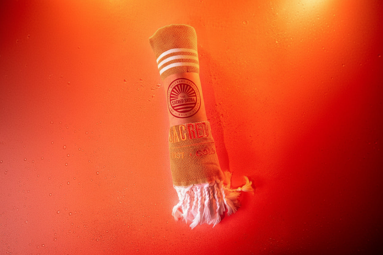 En handduk från The Sacred Sauna, rullad och med logotypen 'THE SACRED SAUNA' samt texten 'HOT · COLD' tryckt på den. Handduken ligger på en fuktig yta med orange bakgrund, vilket skapar en varm och inbjudande atmosfär.